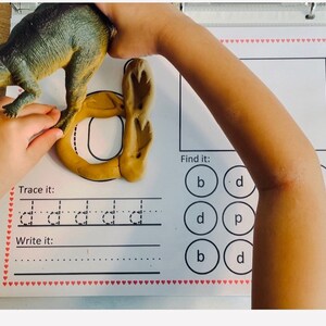 May include: A child's educational worksheet with a brown playdough letter 'd' and a dinosaur toy. The worksheet has instructions to trace and write the letter 'd' and a section to find the letter 'd' in a circle of letters.