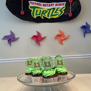 May include: A set of eleven cupcakes decorated with green frosting and topped with a Teenage Mutant Ninja Turtles themed topper. The toppers are green and red and feature the number 11.
