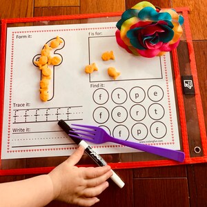 May include: A white learning mat with a red border and hearts printed around the edge. The mat has a letter 'f' formed with goldfish crackers, a section to trace the letter 'f', and a section to write the letter 'f'. There is a section to find the letter 'f' in a grid of letters. The mat is on a wooden surface with a purple fork and a black marker.