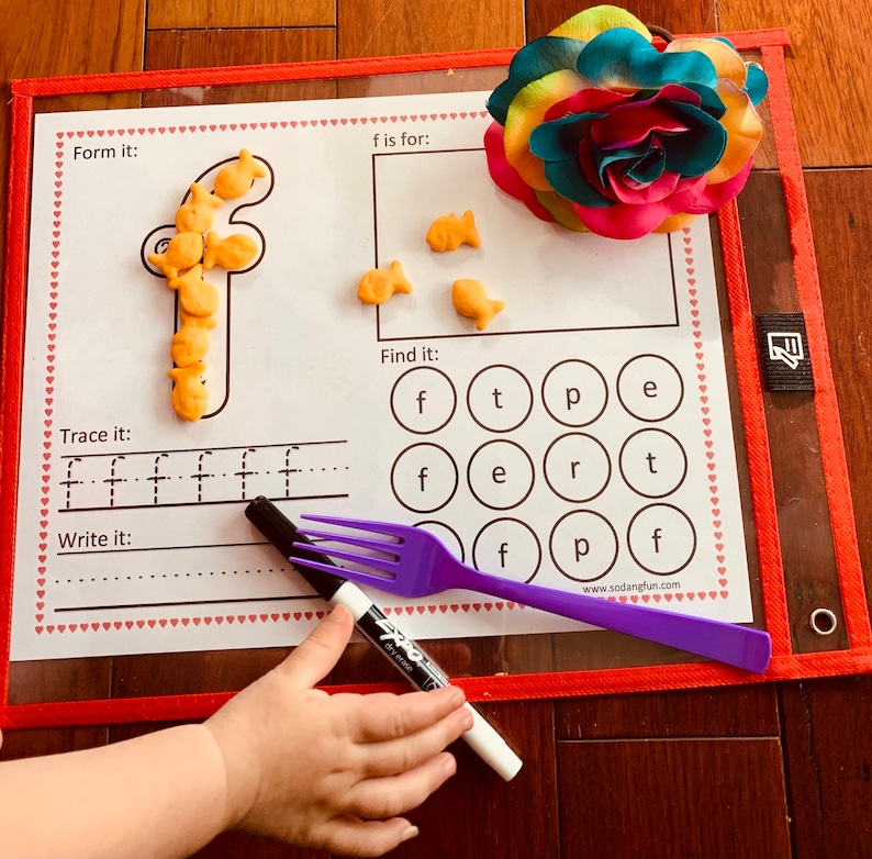 May include: A white learning mat with a red border and hearts printed around the edge. The mat has a letter 'f' formed with goldfish crackers, a section to trace the letter 'f', and a section to write the letter 'f'. There is a section to find the letter 'f' in a grid of letters. The mat is on a wooden surface with a purple fork and a black marker.