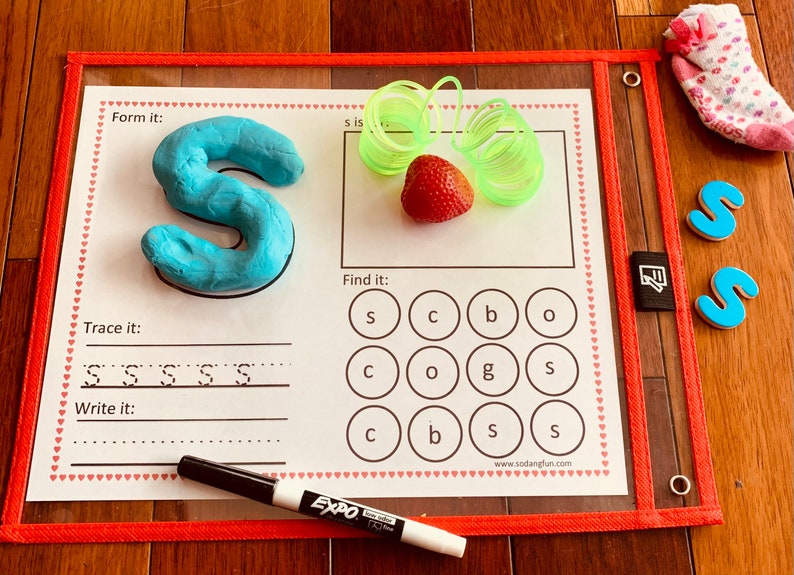 May include: A learning mat with a blue playdough letter S, a red strawberry, green spring toys, and a black marker. The mat has a section to trace the letter S, a section to write the letter S, and a section to find the letter S in a grid of letters.