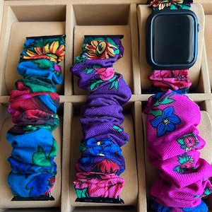 May include: Three colorful patterned watch bands in a wooden box. The bands are made of fabric and have a variety of floral designs. The bands are in blue, purple, and pink.