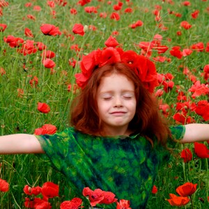 K&ouml;nnte beinhalten: Ein junges M&auml;dchen mit roten Haaren und einer Mohnblumenkrone steht in einem Feld mit roten Mohnblumen und hat die Arme ausgestreckt.