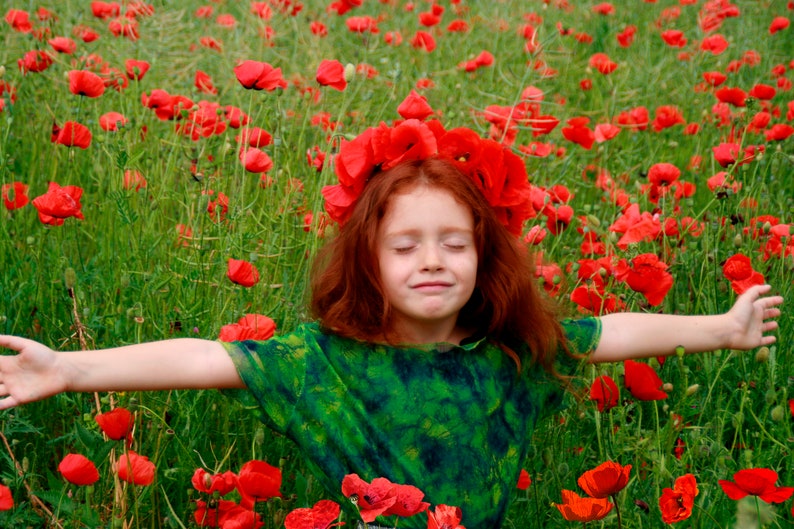 K&ouml;nnte beinhalten: Ein junges M&auml;dchen mit roten Haaren und einer Mohnblumenkrone steht in einem Feld mit roten Mohnblumen und hat die Arme ausgestreckt.