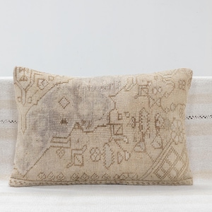 May include: A rectangular throw pillow featuring a woven, textured surface in shades of beige and light brown. The pillow displays a faded, intricate geometric pattern with linear motifs, diamonds, and stylized floral shapes across the front.