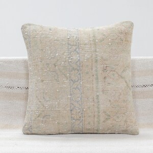 May include: A square throw pillow featuring a woven textile with a faded beige and cream base color. The surface displays a subtle, intricate pattern in muted blue and sage green tones, characterized by worn textures and a weathered appearance.