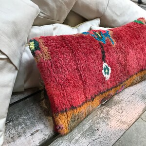 Könnte beinhalten: Ein rotes, orangefarbenes und blaues Kissen mit Muster und Fransenrand. Das Kissen ist aus weichem, gewebtem Stoff und liegt auf einer Holzbank.