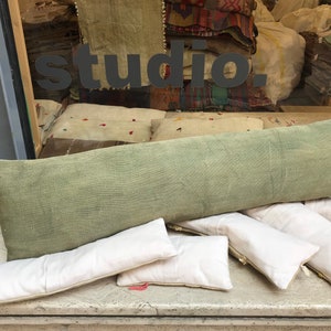 May include: A long green pillow with a white fabric cover, and five smaller white pillows with a white fabric cover. The pillows are arranged on a light brown surface. The word "studio" is visible in the background.