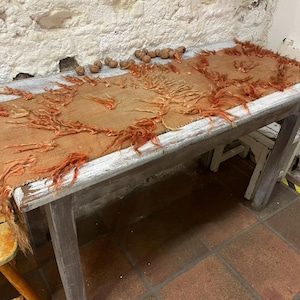 May include: A rustic, rectangular wooden table with a distressed white finish. The table is covered with a tan fabric adorned with orange fringe. Several walnuts are scattered on the fabric. The table is in a room with a stone wall.