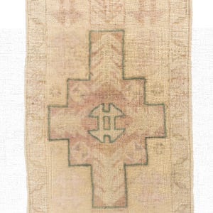 May include: A rectangular, beige rug with a central cross-like design in muted red and green. The rug has a textured appearance with a faded, vintage aesthetic. The edges are finished with a darker, possibly purple, trim.