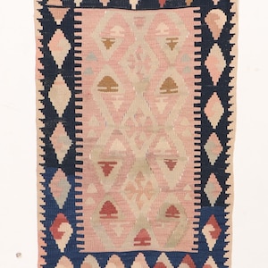 Puede incluir: Una alfombra rectangular, tejida a mano, con un patrón geométrico rosa y beige en el centro. La alfombra está bordeada por un patrón de diamantes azul oscuro. Se incorporan acentos de rojo, beige y azul claro. La alfombra es probablemente de lana.
