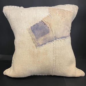 Op de afbeelding: Crèmekleurig vierkant kussen met een patchwork ontwerp. Het kussen heeft een textuurstof en is versierd met lappen vervaagde blauwe en beige stof, aan elkaar gestikt. De algehele esthetiek is rustiek en handgemaakt.