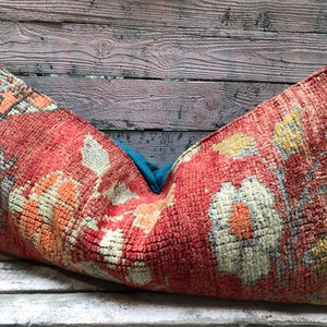 May include: A large, rectangular pillow with a red, orange, and white patterned fabric. The pillow is made from a woven fabric with a textured surface. The pillow is lying on a wooden surface.