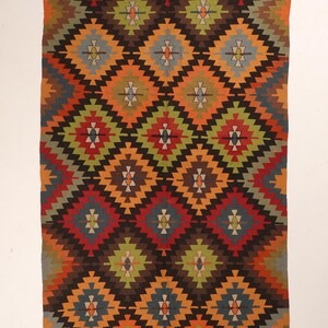 May include: A rectangular, woven rug with a repeating diamond pattern. The design features diamonds in shades of red, orange, green, blue, and brown, set against a dark brown background. The rug has a border with a geometric pattern.