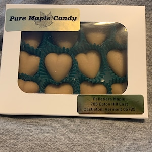 Op de afbeelding: Een witte doos met een transparant plastic venster dat een dozijn hartvormige ahorn snoepjes laat zien. De doos heeft een gouden label met de tekst "Pure Maple Candy" en een kleiner gouden label met de tekst "Pelletiers Maple, 785 Eaton Hill East, Castleton, Vermont 05735".