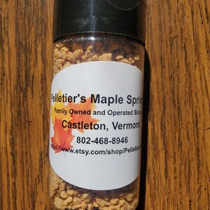 Op de afbeelding: Een doorzichtige plastic kruidenpot met een zwarte deksel, gevuld met kleine, bruinachtige korrels. Het etiket luidt "Pelletier's Maple Springs" en bevat tekst over het bedrijf en een websiteadres. De pot staat op een houten oppervlak.