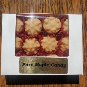 May include: A white box with a clear plastic window showing nine maple candies in red and white paper cups. The box has a gold label that reads "Pure Maple Candy".