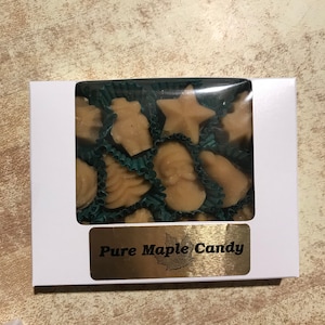 May include: A white box with a clear plastic window showing a variety of maple candy shapes. The candy is in green paper cups. The box has a gold label that says "Pure Maple Candy".