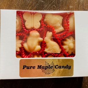 May include: A white box with a clear plastic window showing a variety of maple candies in red and white wrappers. The candies are shaped like butterflies, frogs, rabbits, and other animals. The box has a gold label that says "Pure Maple Candy".