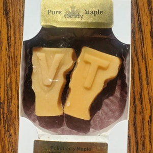 May include: A white box with a gold label that reads "Pure Candy Maple" and a smaller gold label that reads "Pelletier's Maple 785 Eaton Hill East Castleton Vermont 05735". The box contains two maple candy pieces shaped like the letters "V" and "T".