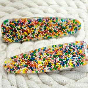 Op de afbeelding: Twee transparante haarclips met kleurrijke sprinkels erin ingebed. De clips zijn ovaalvormig en hebben een gladde, glanzende afwerking.