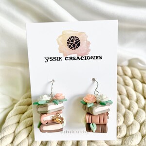 May include: A pair of handmade earrings featuring a stack of books with flowers on top. The earrings are made of polymer clay and have a brown, pink, and green color scheme. The earrings are labeled "YSSIK CREACIONES" and "handmade earrings".