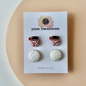 May include: A pair of earrings featuring two brown mugs with white floral designs and two white concha bread earrings. The earrings are displayed on a white card with the text "yssik creaciones" and "Handmade earrings".