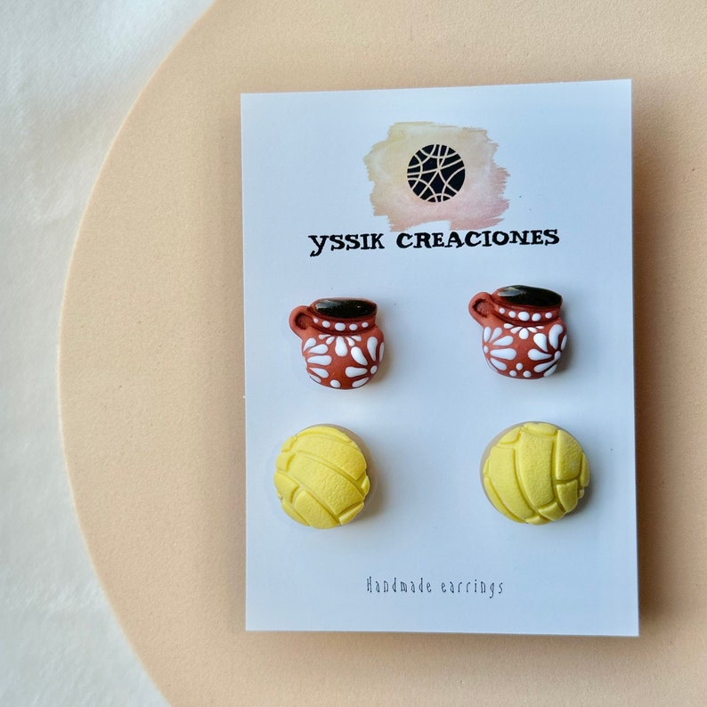 May include: A pair of handmade earrings on a white card. The earrings feature two brown mugs with white floral designs and two yellow concha bread shapes. The card has the text "yssik creaciones" and "Handmade earrings".