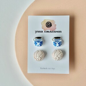 May include: A pair of white earrings shaped like a volleyball and a pair of earrings shaped like a blue and white mug. The earrings are on a white card with the text "yssik creaciones" and "handmade earrings".