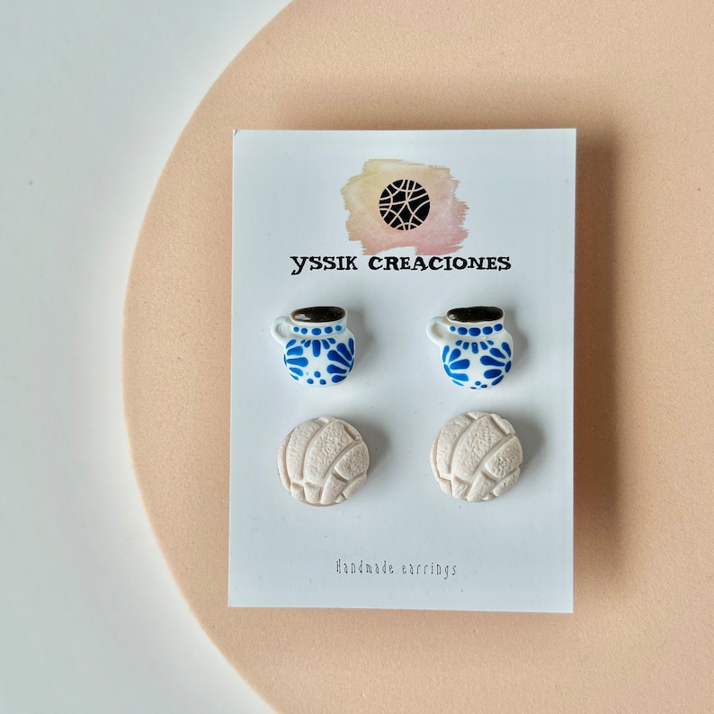 May include: A pair of white earrings shaped like a volleyball and a pair of earrings shaped like a blue and white mug. The earrings are on a white card with the text "yssik creaciones" and "handmade earrings".