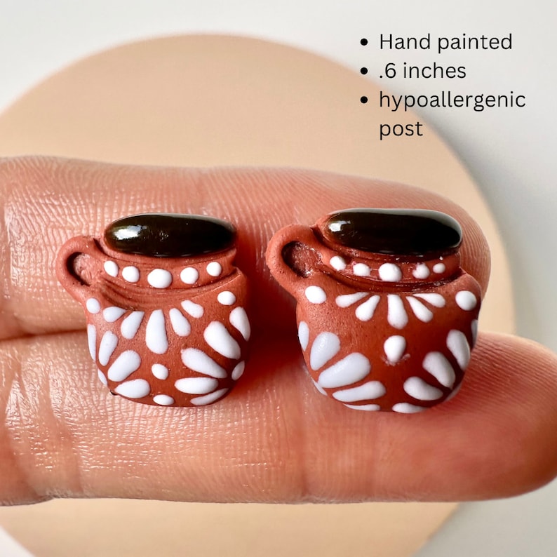 May include: Hand-painted, terracotta-colored earrings shaped like miniature coffee mugs. Each mug features a white floral pattern and a dark brown, glossy top, with a .6-inch size. The earrings have hypoallergenic posts.