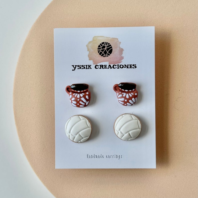 May include: A pair of earrings featuring two brown mugs with white floral designs and two white concha bread earrings. The earrings are displayed on a white card with the text "yssik creaciones" and "Handmade earrings".