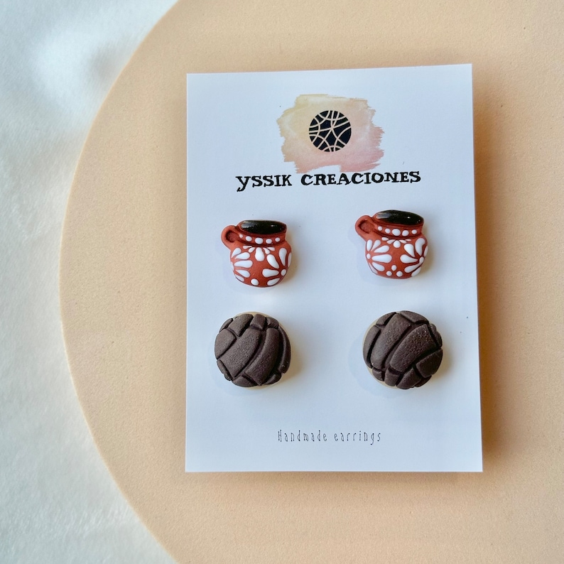 May include: A pair of earrings on a white card. The earrings are in the shape of a brown mug with white floral designs and a dark brown top. Below are earrings shaped like brown concha bread. The card has the text "yssik creaciones" and "Handmade earrings."