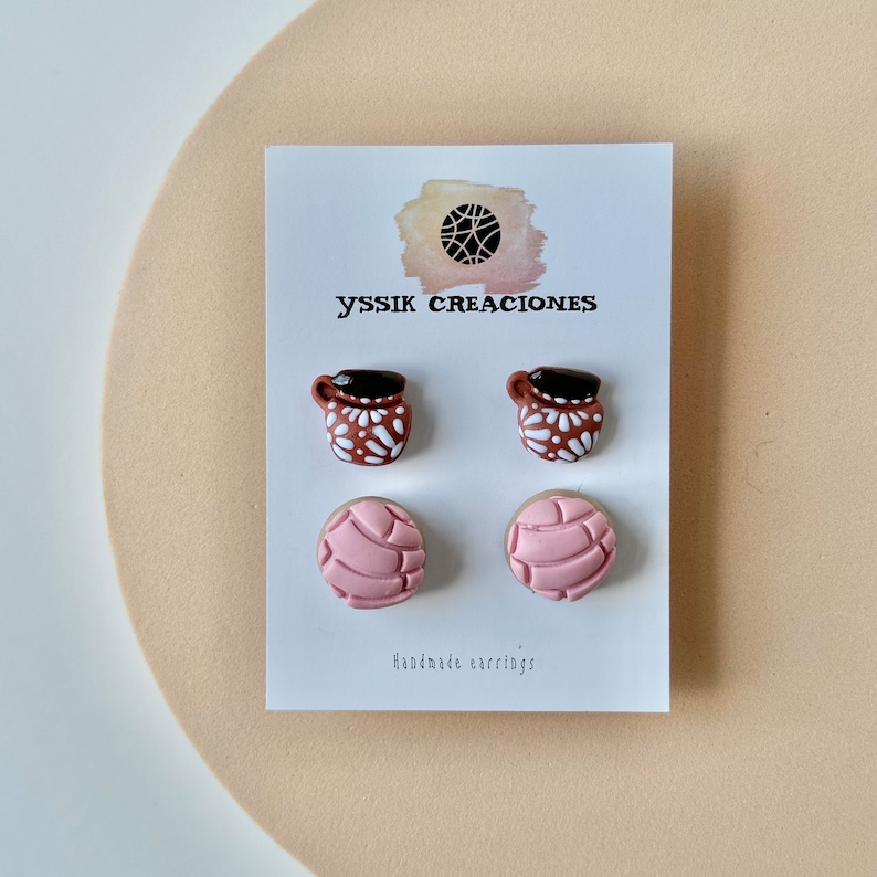 May include: Handmade earrings featuring two pairs of miniature food-themed designs. One pair depicts brown mugs with white floral patterns and black tops. The other pair showcases pink concha bread shapes. The card reads "yssik creaciones" and "Handmade earrings."