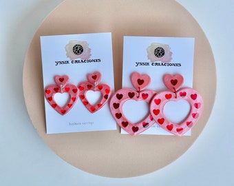 Op de afbeelding: Een paar roze hartvormige oorbellen met rode glitterhartjes. De oorbellen zijn handgemaakt en hebben de tekst "YSSIK CREACIONES" op de kaart.
