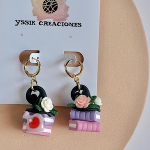 May include: A pair of gold-toned hoop earrings featuring charms shaped like stacks of books topped with black witch hats. One earring has a white rose and a potion bottle with a red heart, while the other has a pink rose. YSSIK CREACIONES text.