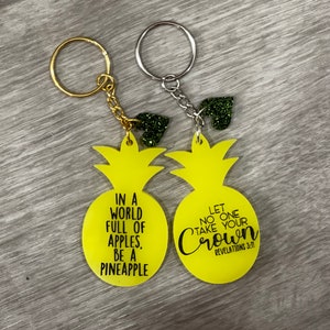 May include: Two yellow pineapple-shaped keychains with black text. One keychain says "In a world full of apples, be a pineapple". The other keychain says "Let no one take your crown, Revelations 3:11".