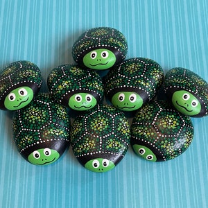 May include: A collection of hand-painted turtle rocks. Each rock features a green turtle face with a smiling expression and a black shell decorated with green and white dots. The rocks are arranged on a blue striped background.
