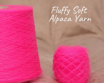 May include: Two bright pink alpaca yarn cones are displayed. The larger cone is upright, while the smaller one is a ball shape. The text "Fluffy Soft Alpaca Yarn" is written in white cursive. The yarn appears soft and fluffy.