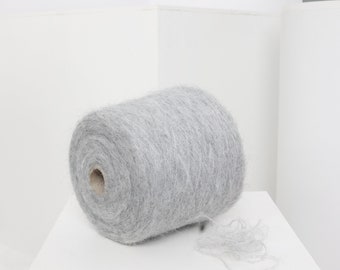 Pode incluir: Um rolo de fio cinza com uma textura felpuda. O fio está enrolado e pronto para ser usado para tricotar ou fazer crochê.