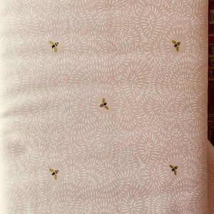 May include: A light pink fabric with a repeating pattern of white dashed lines radiating outwards. Scattered across the fabric are small, detailed illustrations of bees with yellow and black markings. The fabric has a soft, textured appearance.
