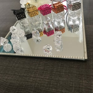 May include: Five decorative glass bottles with metal tops and string hangers. The bottles are filled with clear liquid and are arranged in a row on a mirrored surface with a decorative border. The bottles are in various colours, including black, gold, pink, and silver.