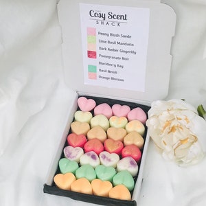May include: A box of heart-shaped wax melts in a variety of colours, including pink, yellow, orange, green, and purple. The box is labelled "The Cosy Scent Shack" and lists the scents of each melt: Peony Blush Suede, Lime Basil Mandarin, Dark Amber Gingerlily, Pomegranate Noir, Blackberry Bay, Basil Neroli, and Orange Blossom.