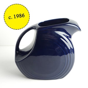 Peut inclure: Un pichet en céramique bleu foncé avec une large ouverture et une poignée incurvée. Le pichet a une finition lisse et brillante et un design légèrement texturé. Le texte "c. 1986" est visible dans le coin supérieur gauche de l'image.