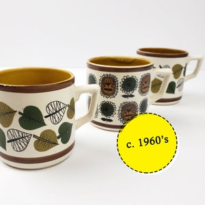 May include: Three vintage ceramic mugs with brown, green and white leaf patterns. The mugs have a yellow interior and a brown rim. The text "c. 1960's" is written on a yellow circle sticker.