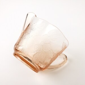 May include: A pink glass cup with a floral design. The cup has a handle and is made of clear glass with a pink tint.