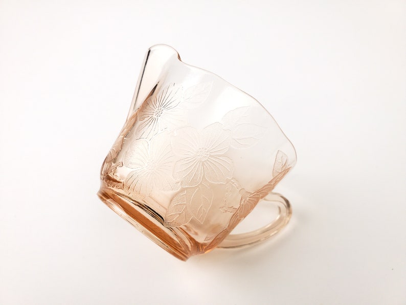 May include: A pink glass cup with a floral design. The cup has a handle and is made of clear glass with a pink tint.