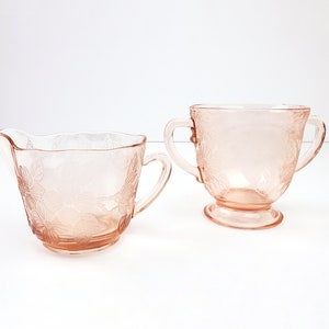 May include: A vintage pink glass creamer and sugar bowl set with a floral design. The creamer has a handle and a pouring spout. The sugar bowl has two handles.