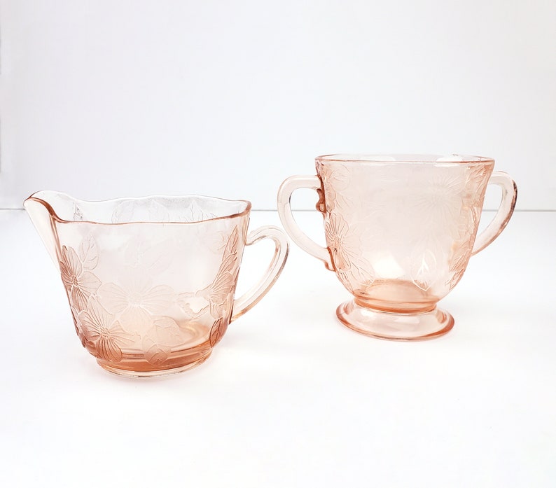 May include: A vintage pink glass creamer and sugar bowl set with a floral design. The creamer has a handle and a pouring spout. The sugar bowl has two handles.