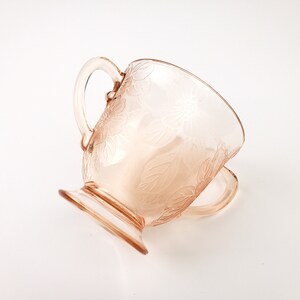 May include: A pink glass creamer with a floral design and a handle. The creamer has a pedestal base.